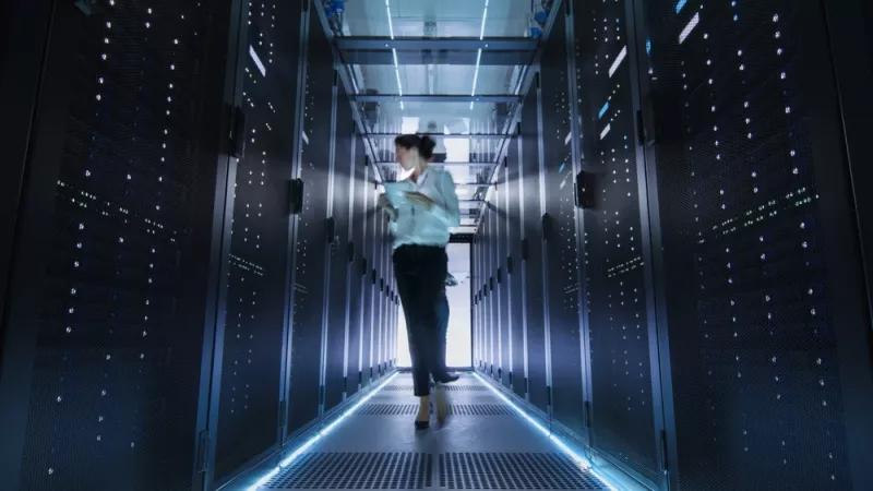 A woman holding a tablet walks down the aisle of a room full of servers.