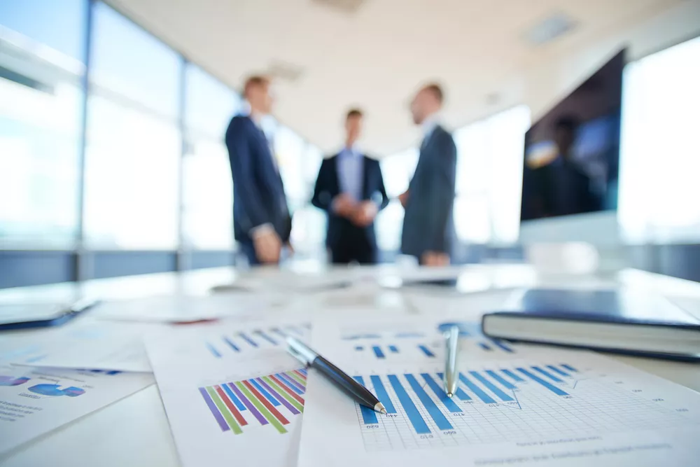 Several documents containing charts and graphs lay on a table in the foreground while three professionals speak to each other in the background.