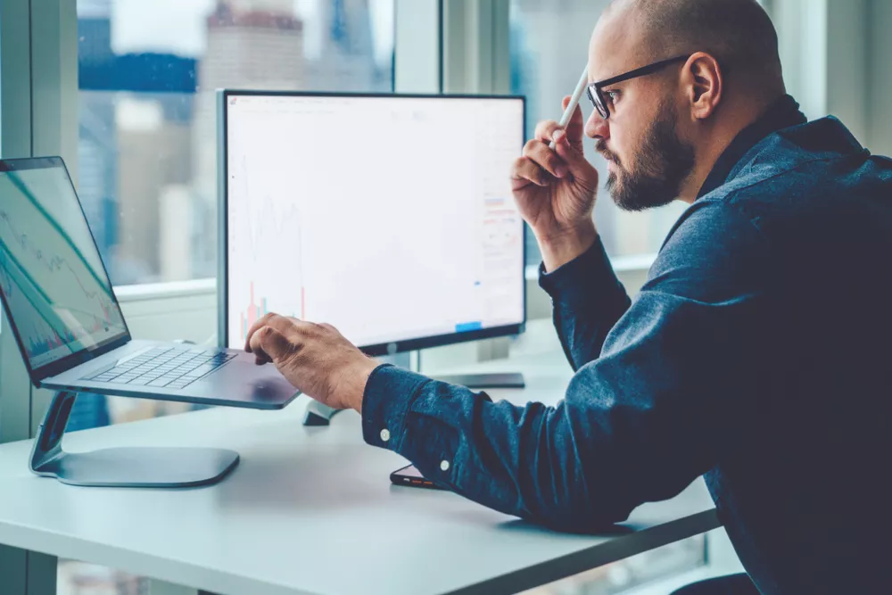 A man sits at a desk with a laptop and a monitor.