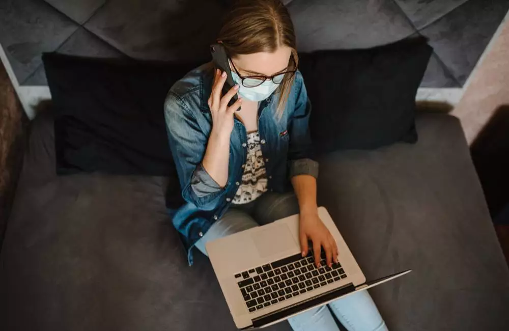 A woman wearing a mask sits with a laptop on her lap as she speaks on the phone.