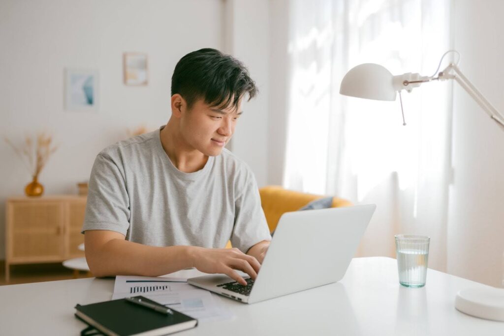 A man in a heather grey tee shirt sits at a table and looks at a laptop computer screen.