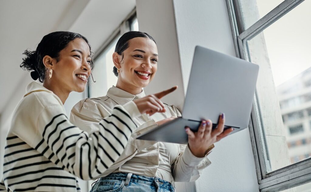 Two smiling, standing women use a laptop; one holds the laptop, the other points to the screen.