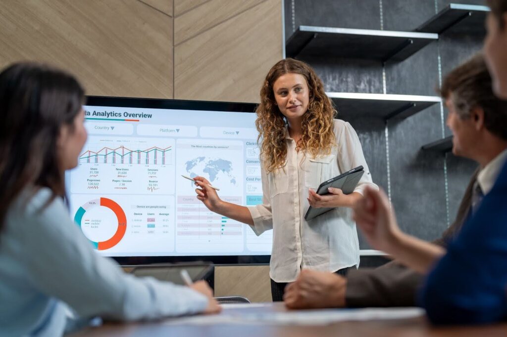 A woman delivers a presentation to a group seated at a conference table; she is pointing to a large screen monitor displaying the title "Data Analytics Overview" and several graphs and maps.