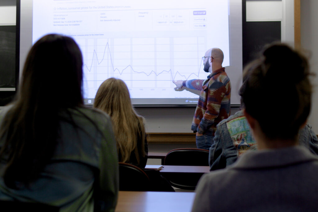 An instructor points to a projected inflation graph while students watch in a classroom setting, focusing on data trends from 1960 to 2023.