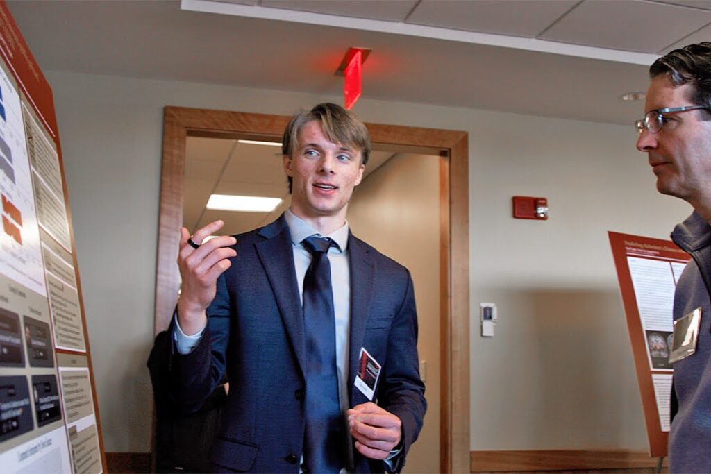 A man in a suit presents research on a poster board to another person.